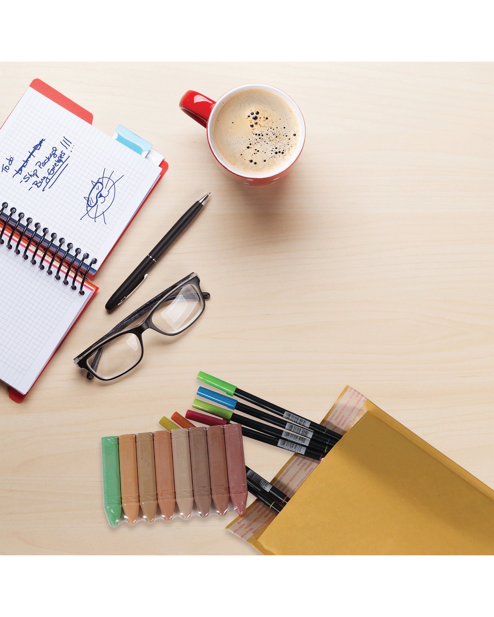 jiffylite mailers displayed with art supplies on a desktop with a notebook, pen, glasses, and cup of coffee